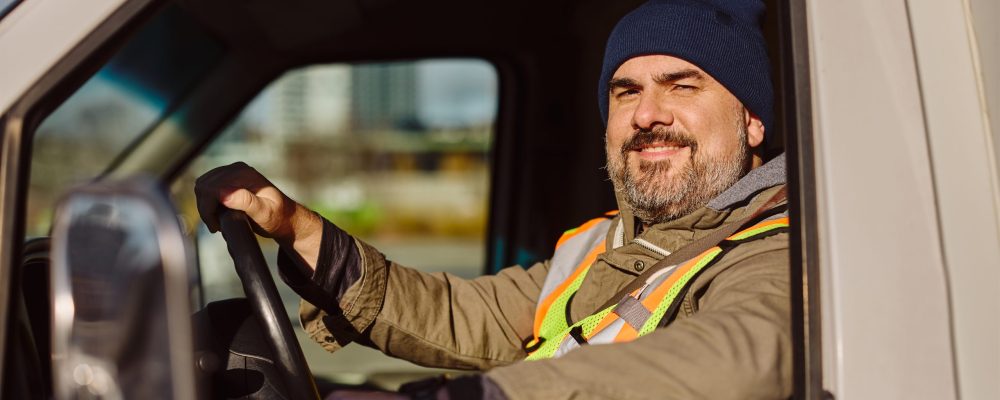 Smiling professional truck driver driving his truck and looking at camera Smiling truck driver looking through side window while driving his truck and looking at camera.